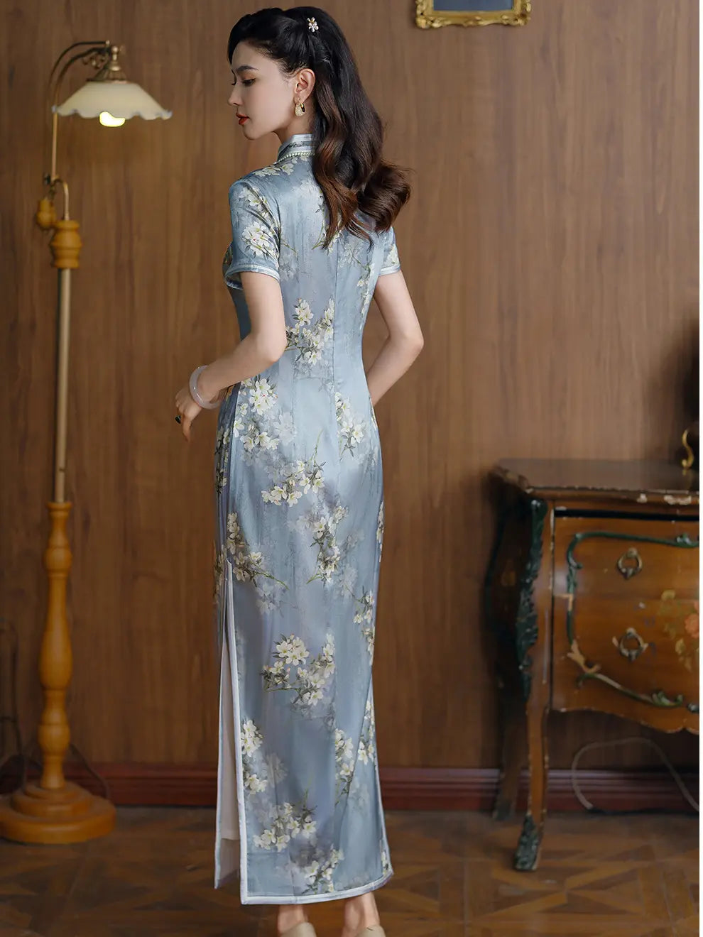 A woman wearing a lLight Blue Mulberry Silk Floral Qipao  cheongsam dress, standing in a vintage room with wood-paneled walls, a floor lamp, and an antique wooden cabinet.