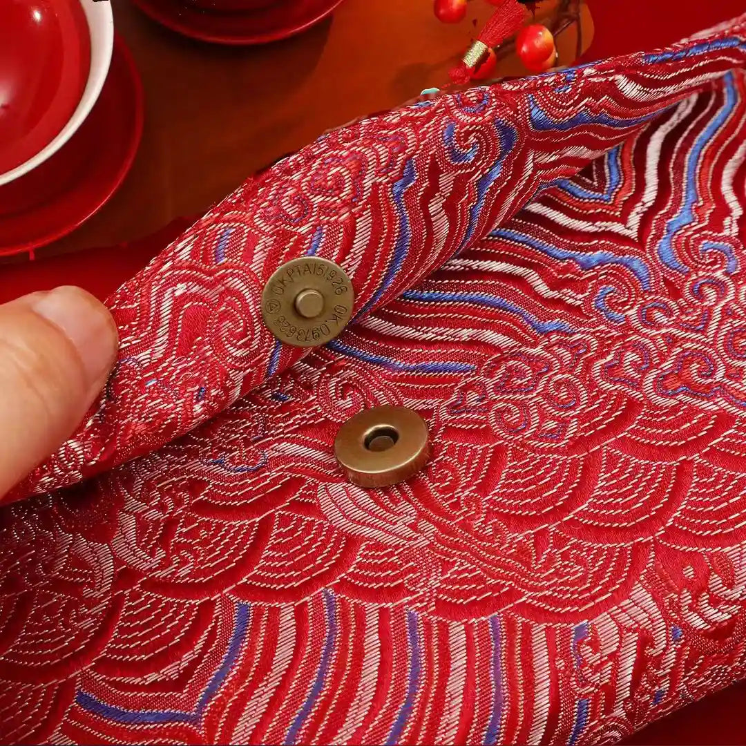 A close-up of a red embroidered fabric Chinese brocade red envelope with a hand lifting one corner to show the interior snap closure. The fabric features intricate wave and cloud patterns in red, white, and blue, with two bronze metal snaps.
