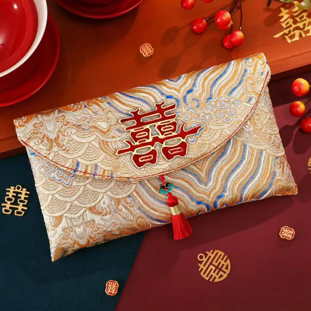 A decorative envelope-style pouch with a gold and blue patterned brocade-like surface, a large red Chinese character 福 on the front, and a red tassel charm, styled on a wooden table with red dishes, berries, and gold double-happiness symbols.