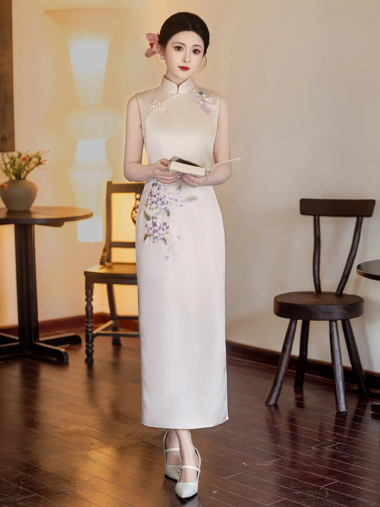 Woman in a long, white Luxury Embroidered New Chinese Style Sleeveless Qipao Dress with floral patterns standing in a room with wooden floors and chairs.