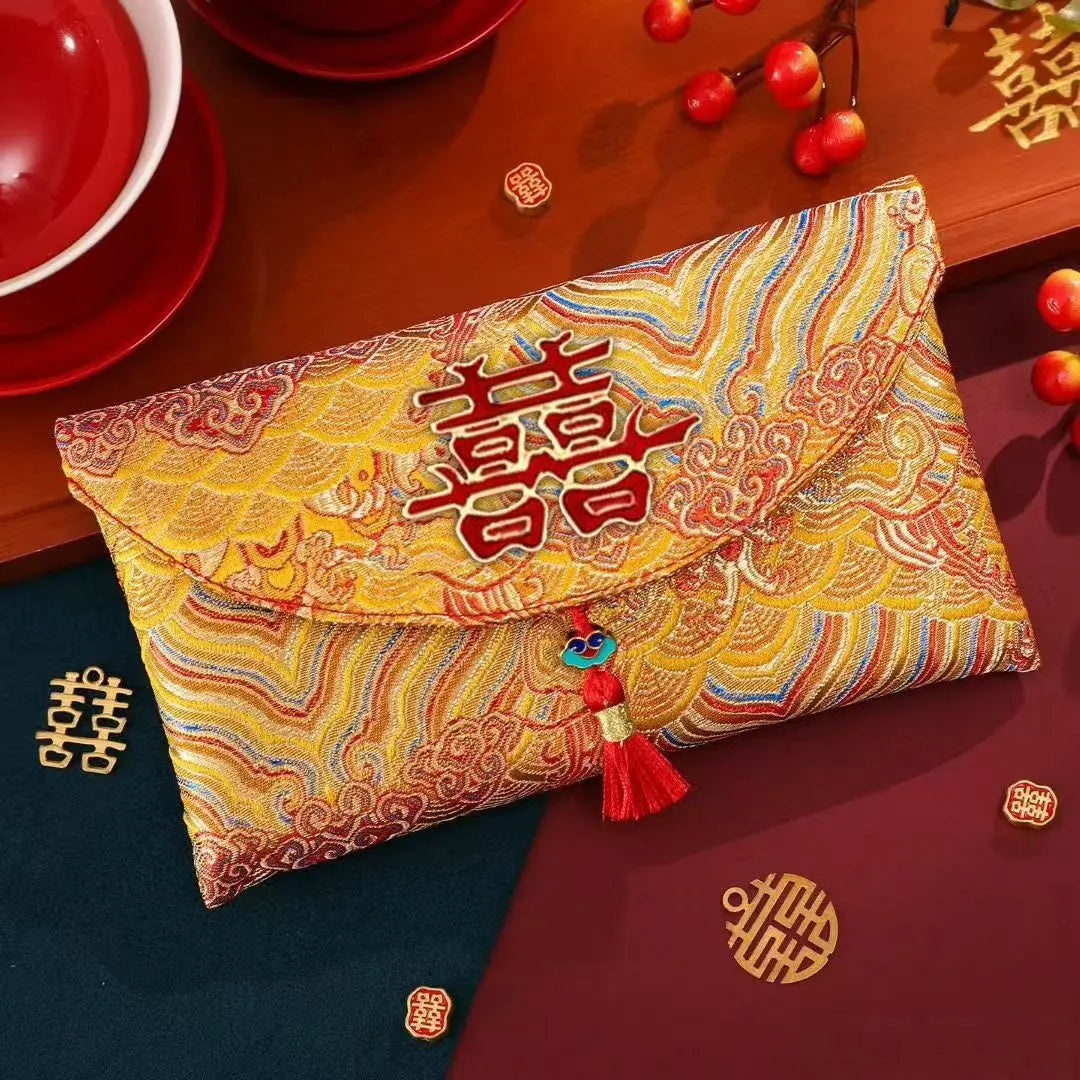 A Chinese wedding envelope clutch bag with a golden-yellow brocade pattern, featuring a large red double happiness symbol on the flap and a red tassel charm, displayed on a dark wooden table with red and dark blue elements.