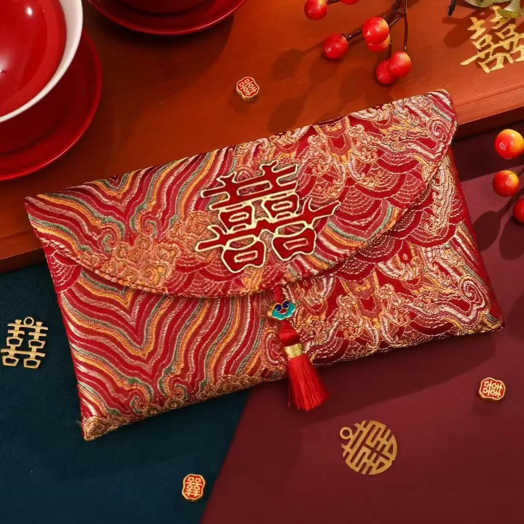 A red and gold embroidered envelope-style clutch with Chinese double-happiness symbols, displayed on a wooden surface with festive props including red cups, berries, and gold decorative pieces.