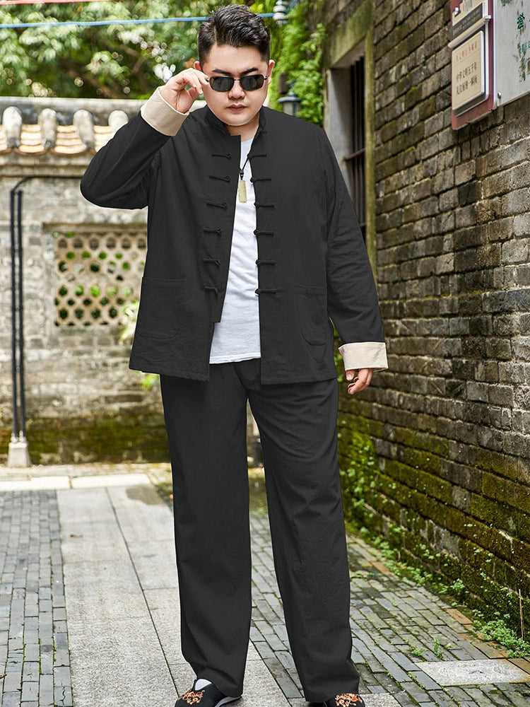 A man wearing a black traditional plus size Chinese-style jacket and matching pants over a white t-shirt, standing on a stone pathway next to a brick wall.