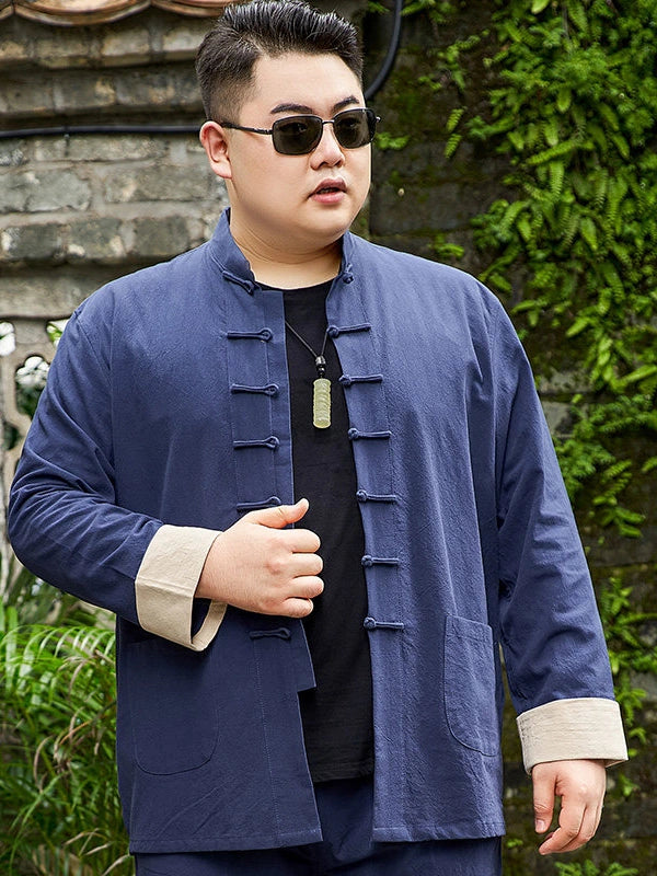 A man wearing a navy blue traditional Chinese-style jacket with beige cuffs and a black shirt underneath, standing outdoors against a stone wall with green foliage.