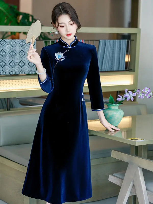 A woman wearing a long navy blue velvet A line cheongsam-style dress with white piping and a floral embroidery near the chest, holding a round fan in a modern indoor setting.