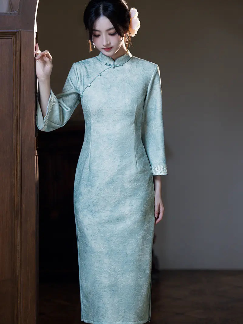 A woman wearing a light sage green cheongsam dress with a subtle textured pattern, standing in a dimly lit room next to a wooden door.