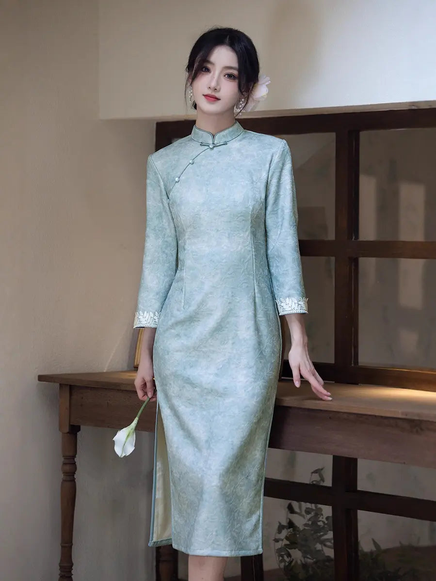 A woman wearing a light sage green cheongsam dress with white lace trim on the cuffs, standing in a warm-toned indoor setting next to a wooden table.
