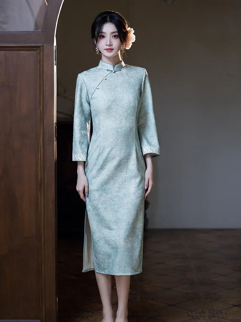 A woman wearing a light sage green textured cheongsam (qipao) dress with three-quarter sleeves and a high collar, standing in a doorway.