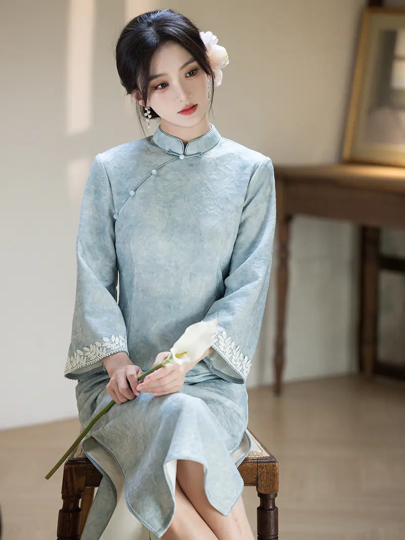 A woman wearing a light blue traditional Chinese cheongsam dress with white lace trim on the cuffs, sitting on a wooden chair in a softly lit indoor setting.