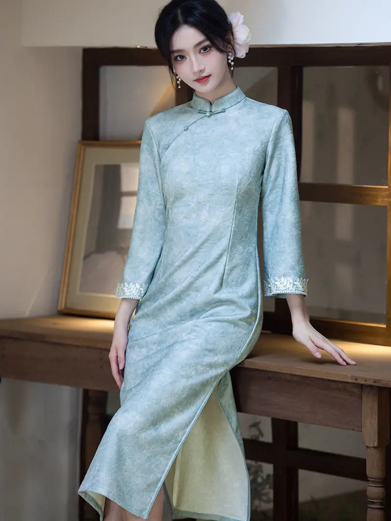 A woman wearing a light sage green Chinese cheongsam (qipao) dress with three-quarter sleeves and a high collar, leaning against a wooden table in a warm-toned indoor setting.