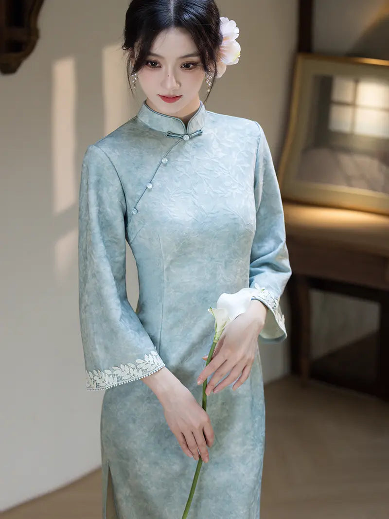 A woman wearing a light blue-green textured cheongsam with white lace trim at the cuffs, holding a white calla lily in a softly lit indoor setting.
