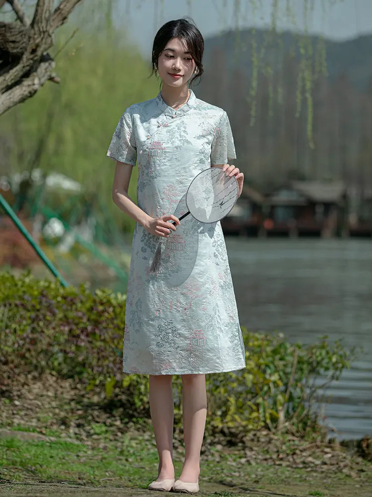 Woman in a light Mint Green Scenic Brocade Loose A Line Qipao Dress holding a fan outdoors with greenery and water in the background