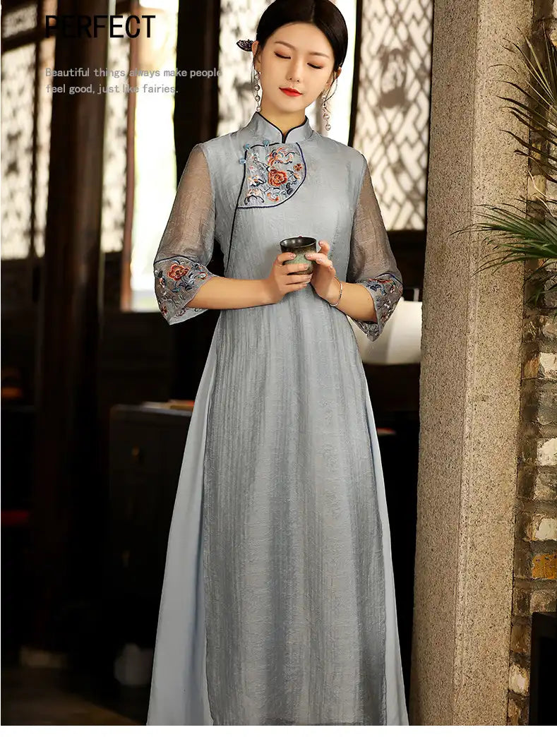 Woman in a traditional light blue Modern A Line Loose Cheongsam Qipao Dress with Sheer Sleeve  holding a cup, standing against a blurred indoor background.