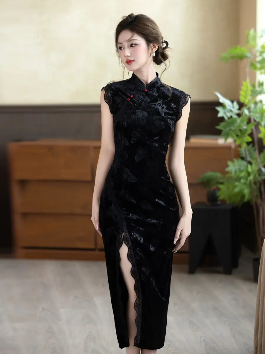A woman modeling a black velvet cheongsam dress with floral embroidery and a high slit, standing in an indoor setting with wooden furniture and a plant in the background.