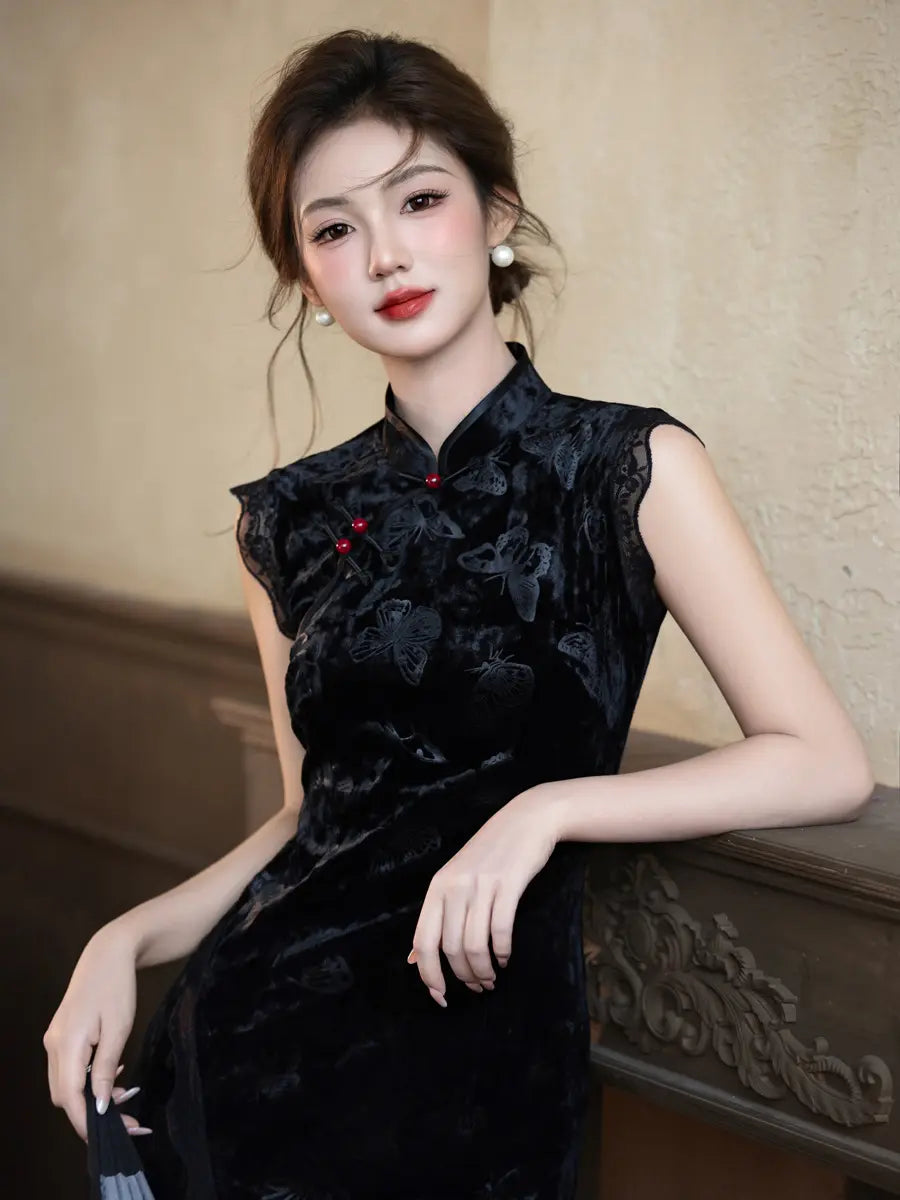 A woman wearing a sleeveless black velvet modern cheongsam-style dress with tonal floral embroidery and small red button accents, posed indoors against a beige wall and carved wooden railing.