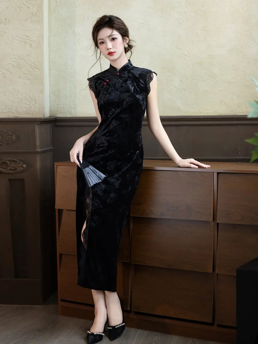 A woman models a black velvet qipao cheongsam dress with a subtle floral jacquard pattern, standing in a room with a wooden cabinet and textured beige wall.