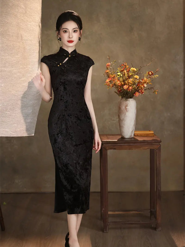 A woman models a black, sleeveless, high-neck, knee-length cheongsam (qipao) dress with a subtle jacquard texture. She is standing in a studio setting with a textured brown backdrop, a wooden table holding a vase of orange flowers, and a beige fabric panel on the left.