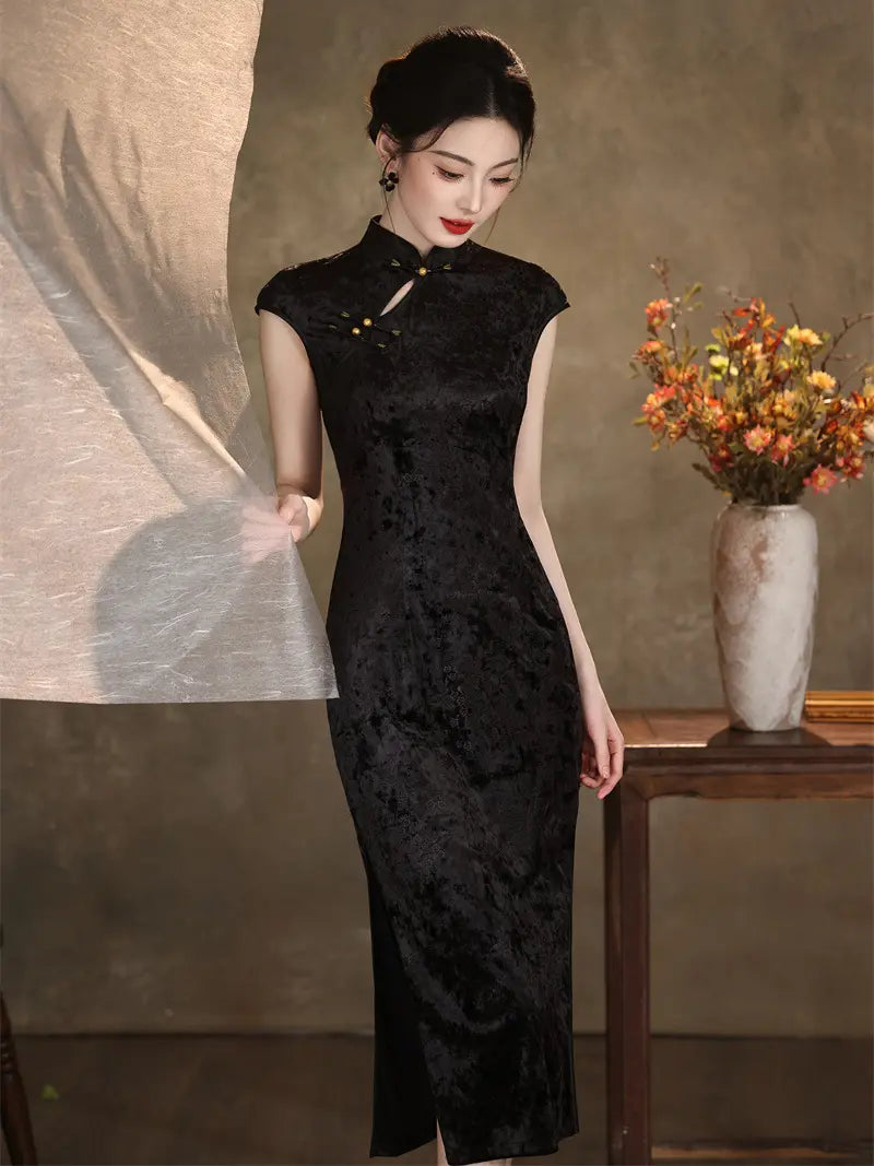 A woman models a black textured Velvet cheongsam/qipao dress with a high collar and three-quarter sleeves, standing in a styled studio setting with a draped fabric panel and a vase of flowers.