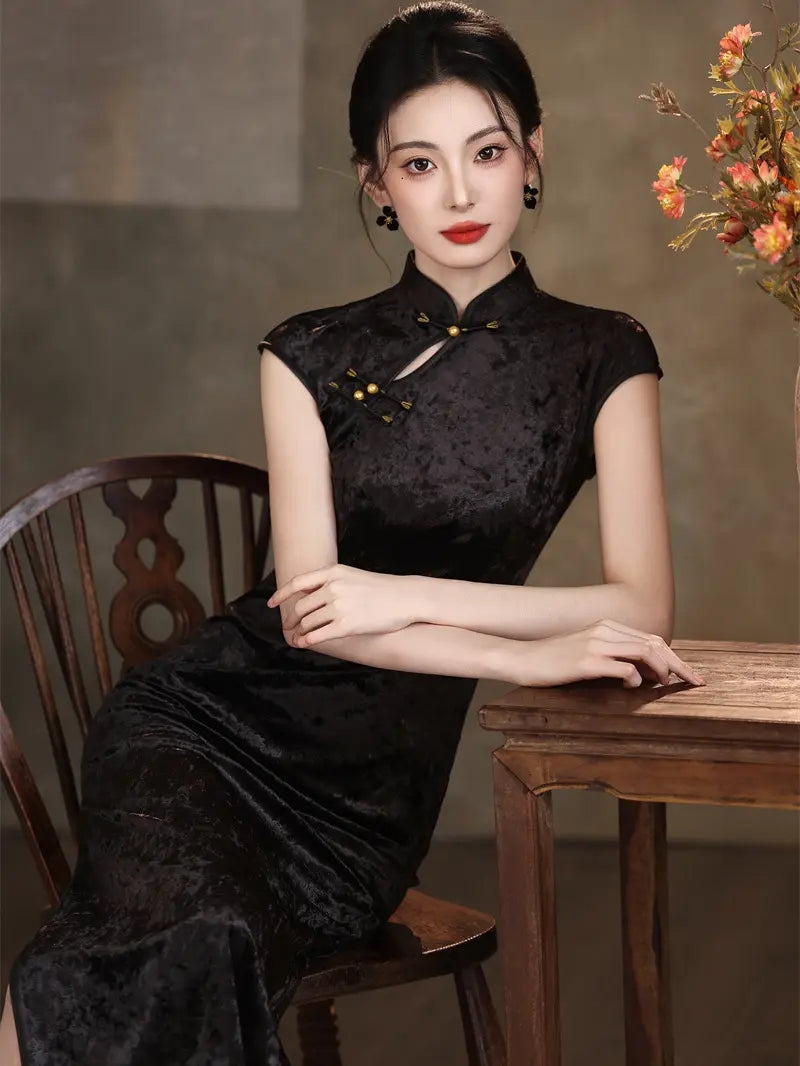 A woman wearing a black textured cheongsam dress sits on a wooden chair next to a wooden table in a styled indoor setting.