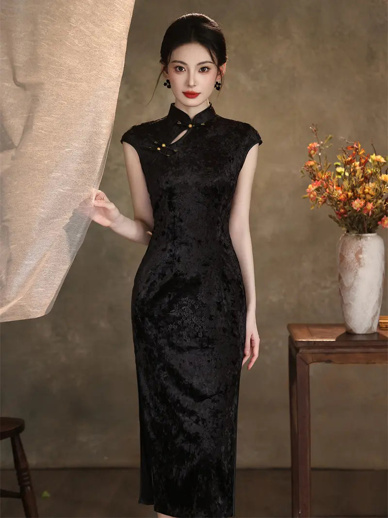 A woman models a black textured cheongsam dress in a styled studio setting with a neutral textured wall, a draped fabric panel, and a side table with dried flowers.