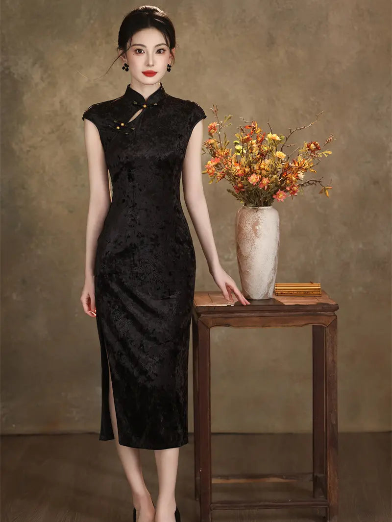 A woman models a black textured cheongsam midi dress with a high slit, standing next to a small wooden table holding a vase of autumn flowers against a textured brown studio backdrop.