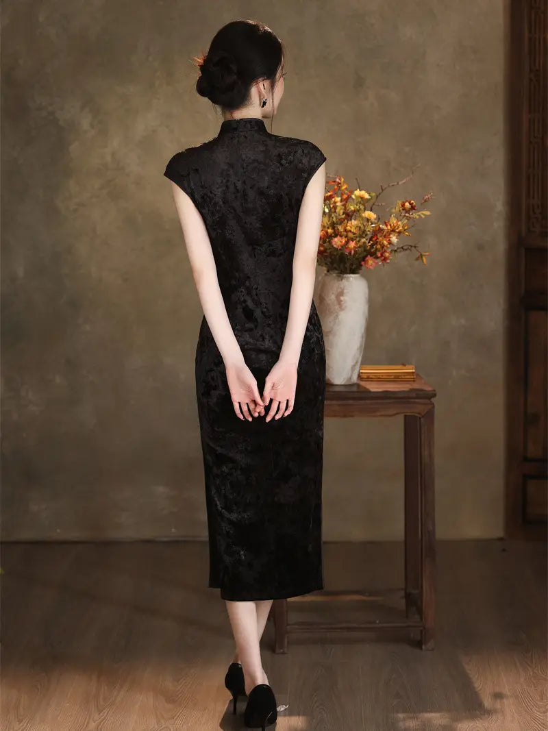 A woman stands with her back to the camera wearing a black textured cheongsam dress, posed in front of a rustic brown wall next to a small wooden table with a vase of autumn flowers.