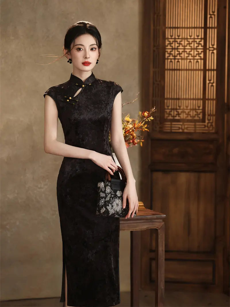 A woman stands indoors wearing a fitted black satin cheongsam-style dress with a floral embroidered pattern near the hem and a small black handbag. The setting includes a textured warm beige wall, a wooden screen, and a wooden table.