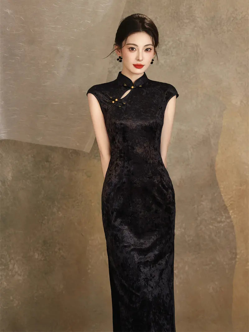 A woman wearing a sleeveless black textured cheongsam-style dress with a high collar and front slit, standing against a textured warm brown backdrop.