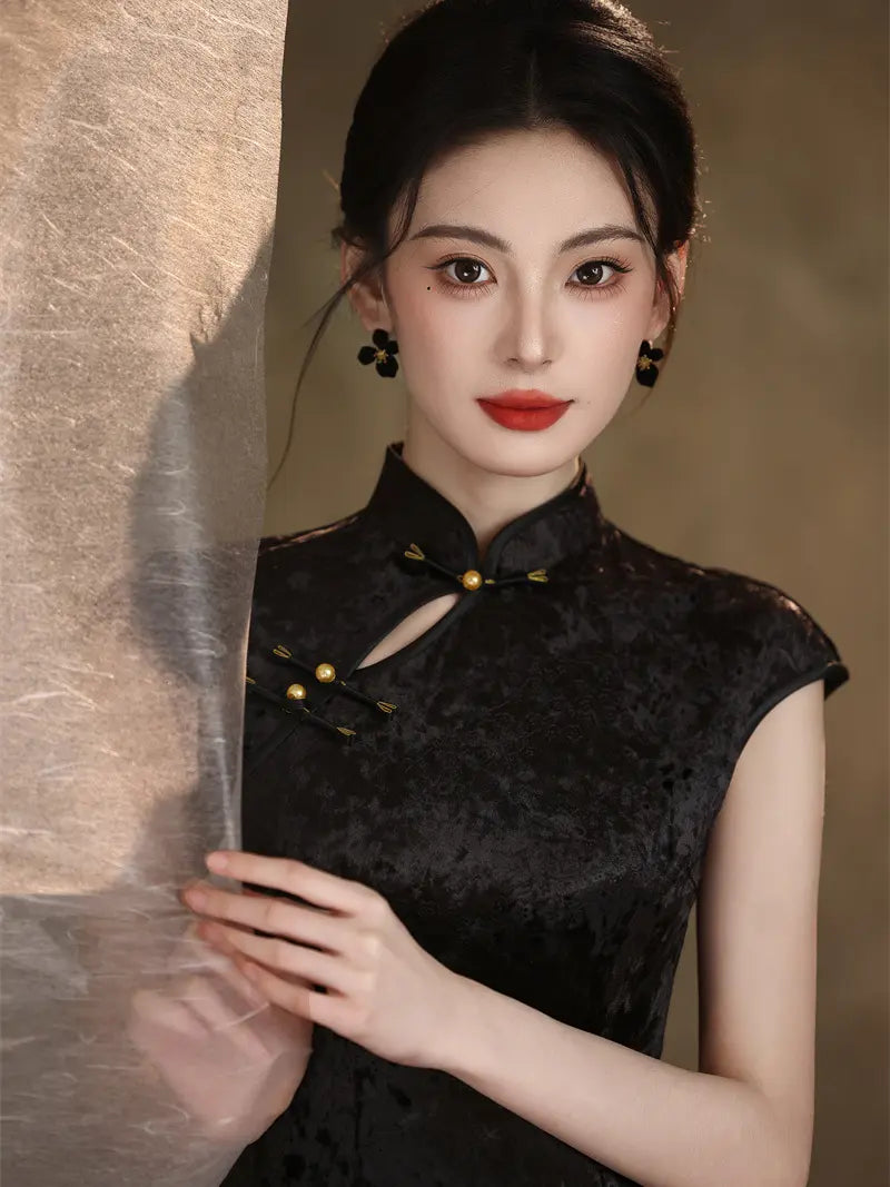 A woman wearing a black textured cheongsam-style sleeveless top with gold-tone buttons and a keyhole neckline, posed against a warm beige backdrop with sheer fabric draped beside her.