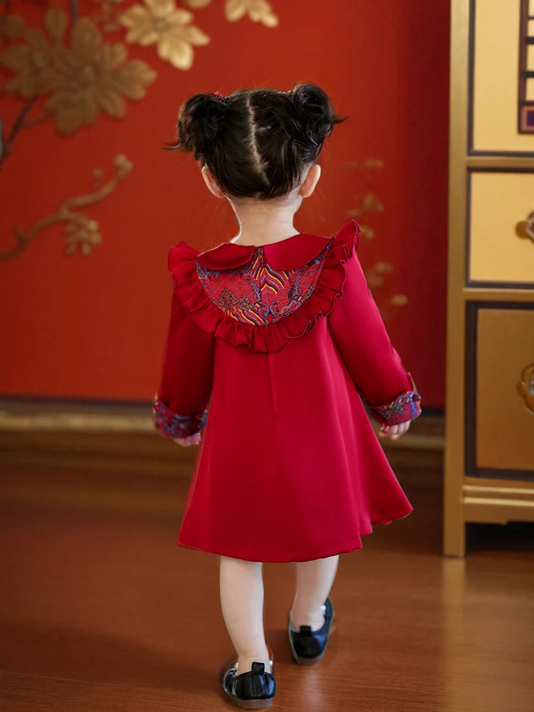 Child wearing a red Modern Chinese Baby Qipao Cheongsam Dress with Tassel  with floral patterns in a room with red walls and gold decorations.