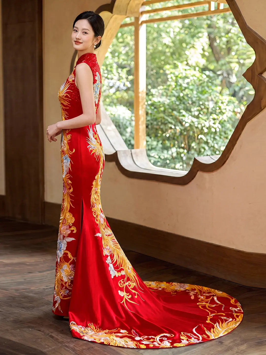 A woman wearing a red and gold Modern Chinese  Sleeveless Phoenix Wedding Qipao with Train  gown with floral embroidery, standing in a room with a decorative window and green foliage outside.