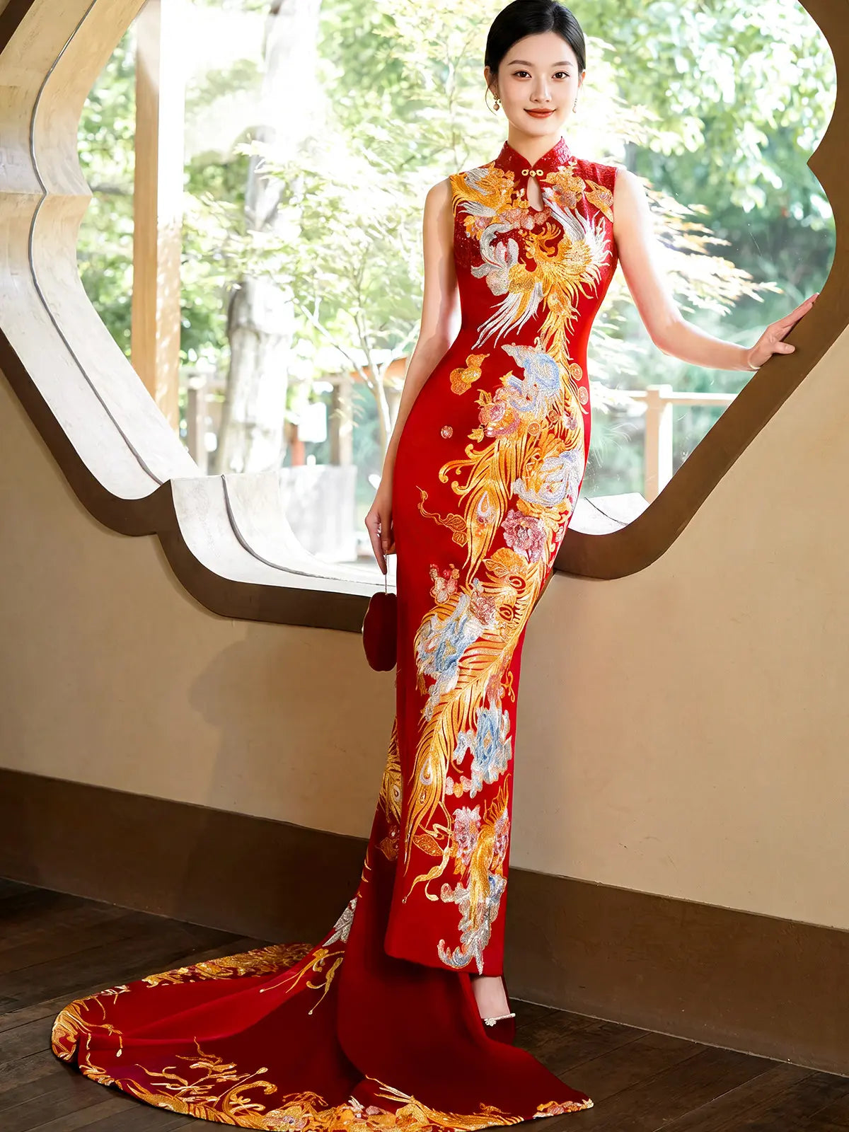 A woman wearing a red sleeveless cheongsam-style wedding gown with large gold, orange, and white phoenix and floral embroidery, standing indoors near a decorative window opening with greenery outside.