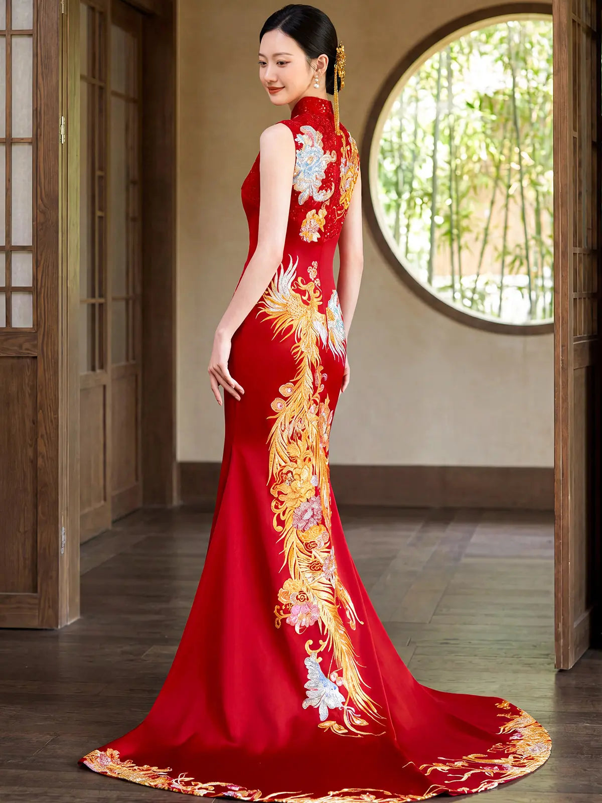A woman wearing a red sleeveless cheongsam-style phoenix Wedding Qipao gown with large gold and white floral phoenix embroidery, standing in a traditional interior with wooden doors and a round window.