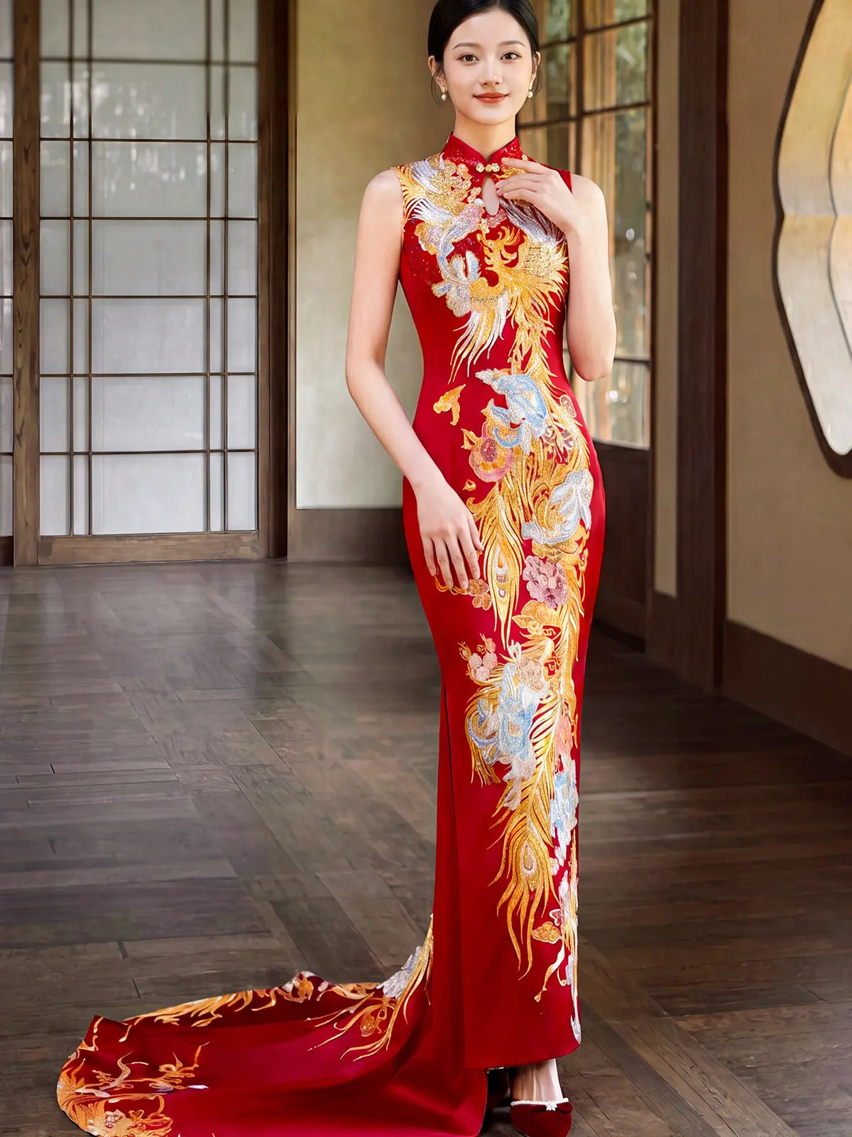 A woman models a sleeveless red cheongsam-style floor-length gown with ornate gold, white, and blue phoenix embroidery and train, standing in a traditional interior with wooden floors and shoji-style doors.