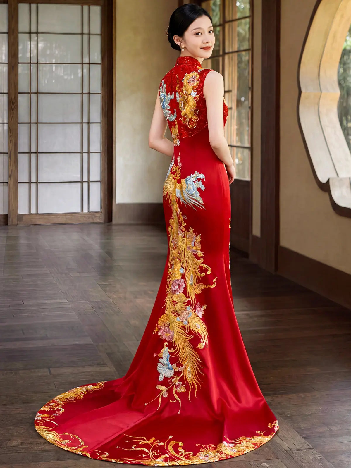 A woman wearing a red Chinese cheongsam/qipao gown with gold and multicolor phoenix embroidery, photographed indoors in a traditional-style room with wooden floors and sliding doors.