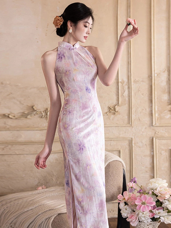 A woman wearing a sleeveless, halter neck cheongsam dress with a light pink and lavender watercolor floral pattern, standing in an elegant room with beige walls and a floral arrangement.