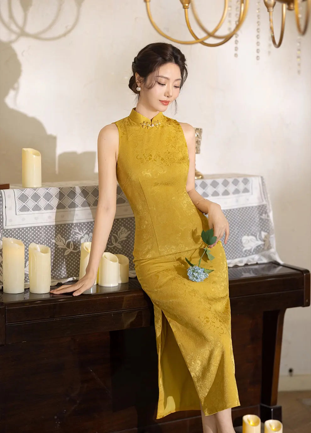 Woman in a Modern Imperial Gold Open Back Jacquard Qipao Dress sitting at a table with candles and a decorative background