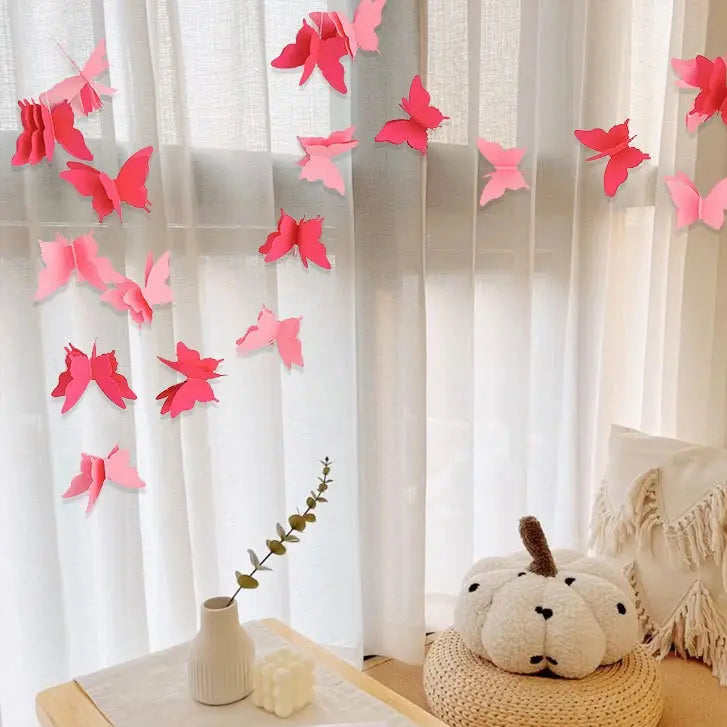 Pink and red paper butterfly wall decorations hanging in a row on a white curtain, styled in a cozy room setting with a small vase, candle, and a plush cream pumpkin on a woven cushion.