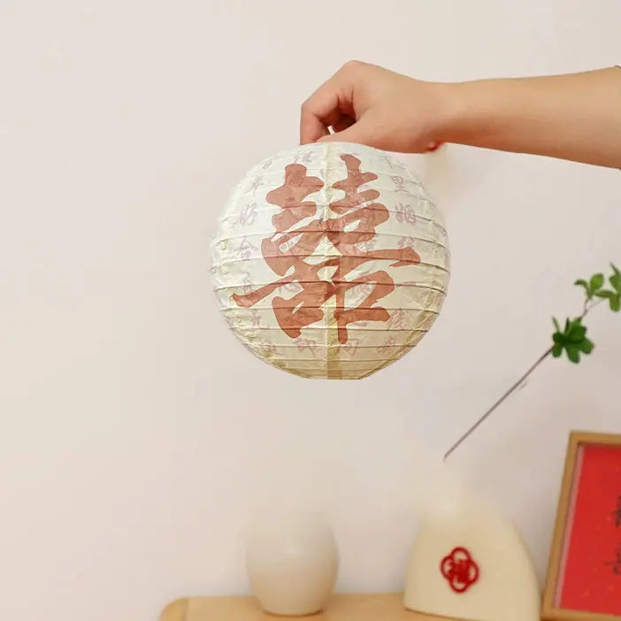 A hand holding a round white paper lantern with a large red Chinese character double happiness printed on it, set against a light beige wall with decorative props in the background.
