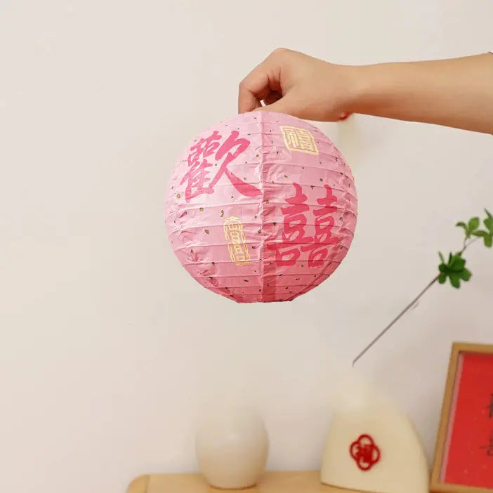 A hand holding a pink paper lantern decorated with red Chinese double happiness characters, set against a warm off-white background with decorative props.