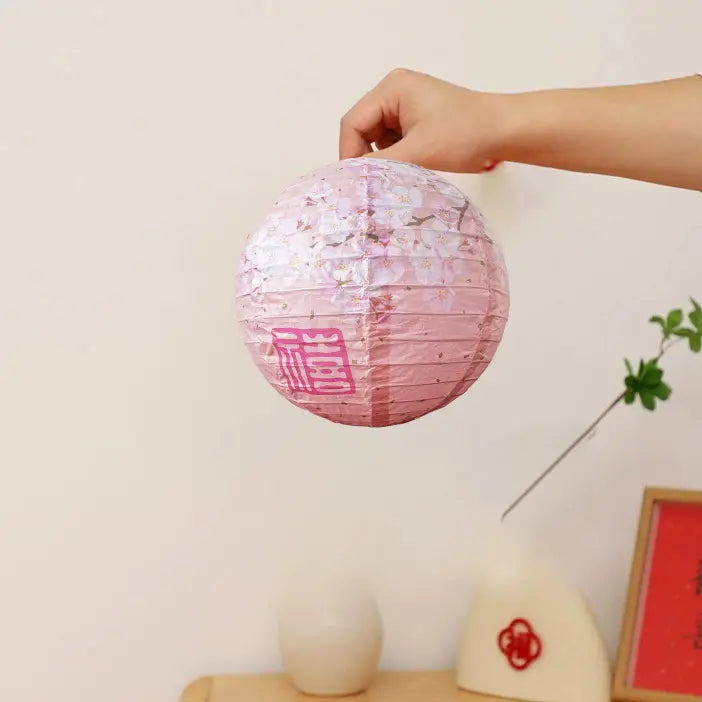 A hand holding a round pink paper lantern decorated with cherry blossom patterns and a red Chinese character, set against a soft beige background with decorative props.