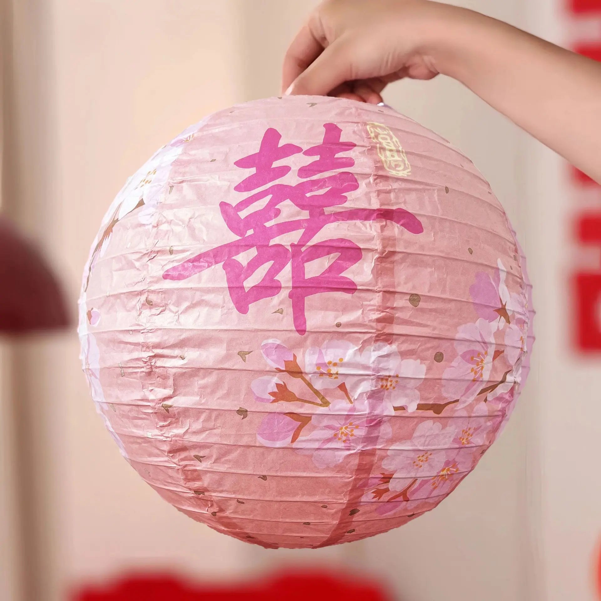 A pink paper lantern with a large pink Chinese double happiness character and cherry blossom illustrations, held by a hand against a softly blurred indoor background.