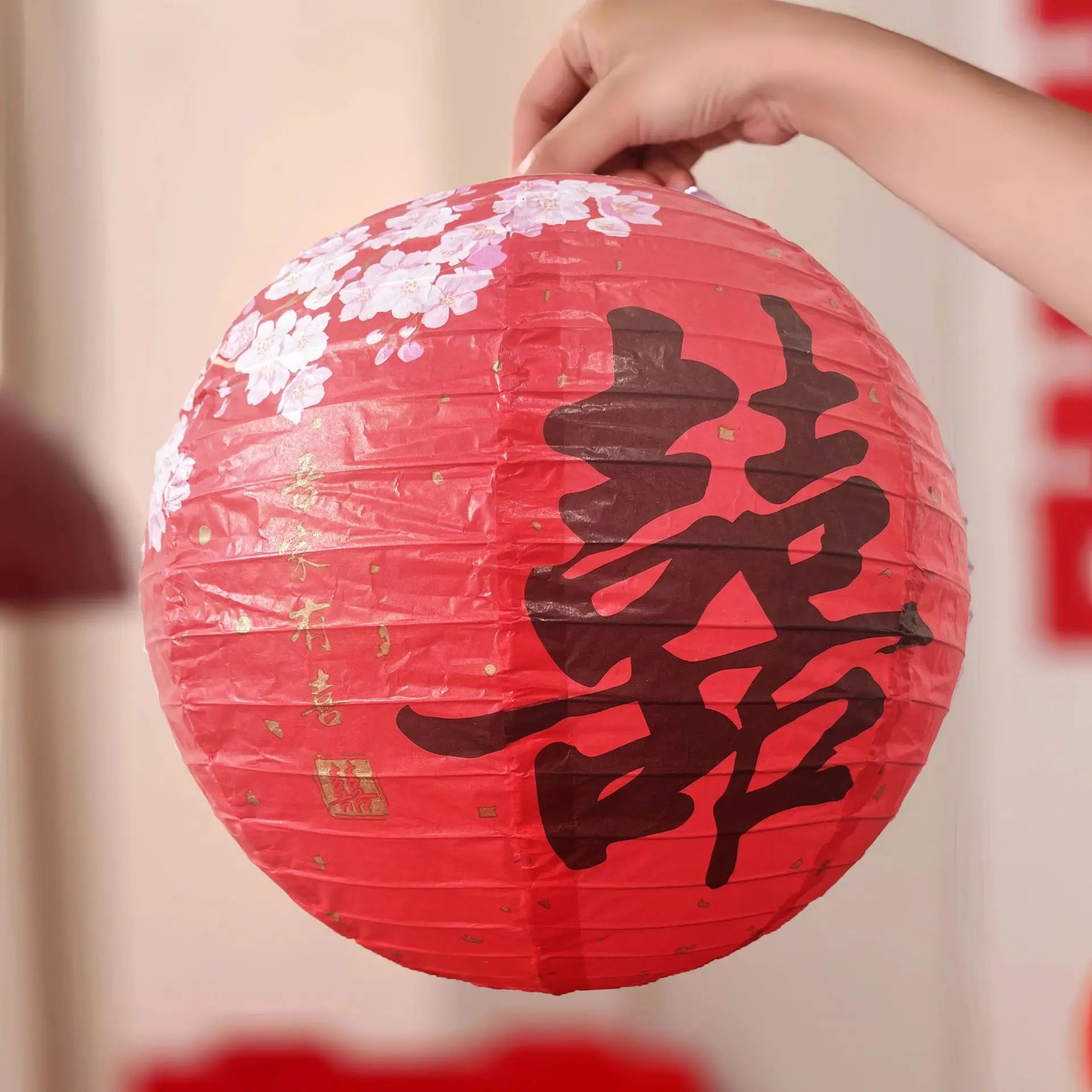 A hand holding a round red paper lantern decorated with white cherry blossoms and a large black Chinese double happiness symbol, set against a softly blurred indoor background.