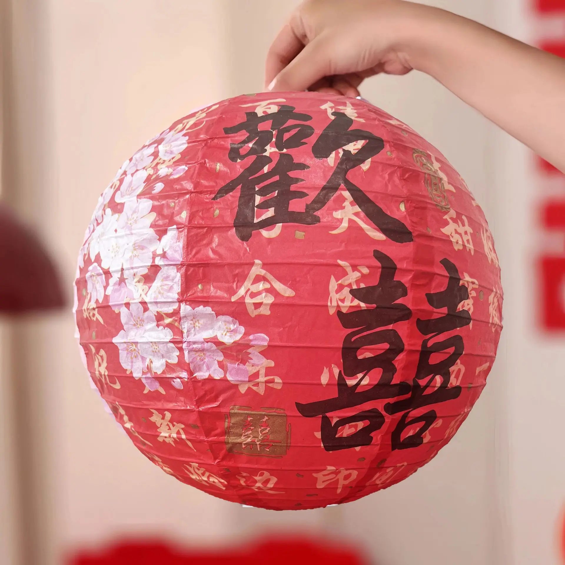 A red round paper lantern with black Chinese calligraphy characters double happiness and white floral patterns, held by a hand against a softly blurred indoor background.