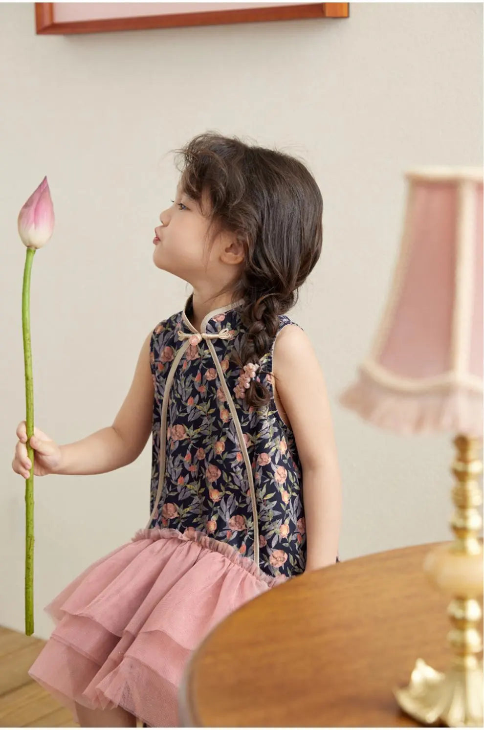 Young girl in a Modern Pink Floral Tulle Qipao Dress  holding a pink flower, with a lamp and wooden surface in the background.