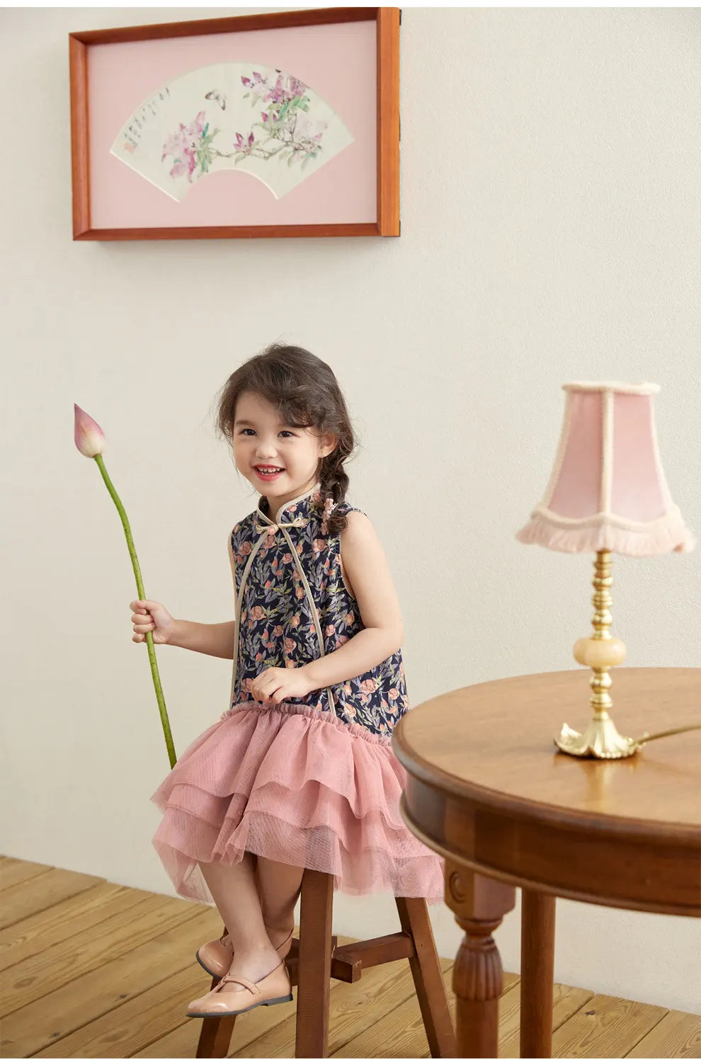 Young girl in a Modern Pink Floral Tulle Qipao Dress  holding a flower, standing on a stool in a room with a lamp and framed picture.