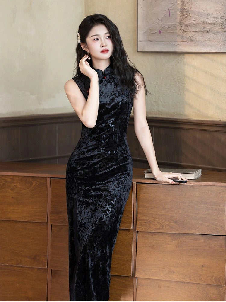 A woman wearing a sleeveless black velvet cheongsam qipao maxi dress, posing indoors next to a wooden cabinet.