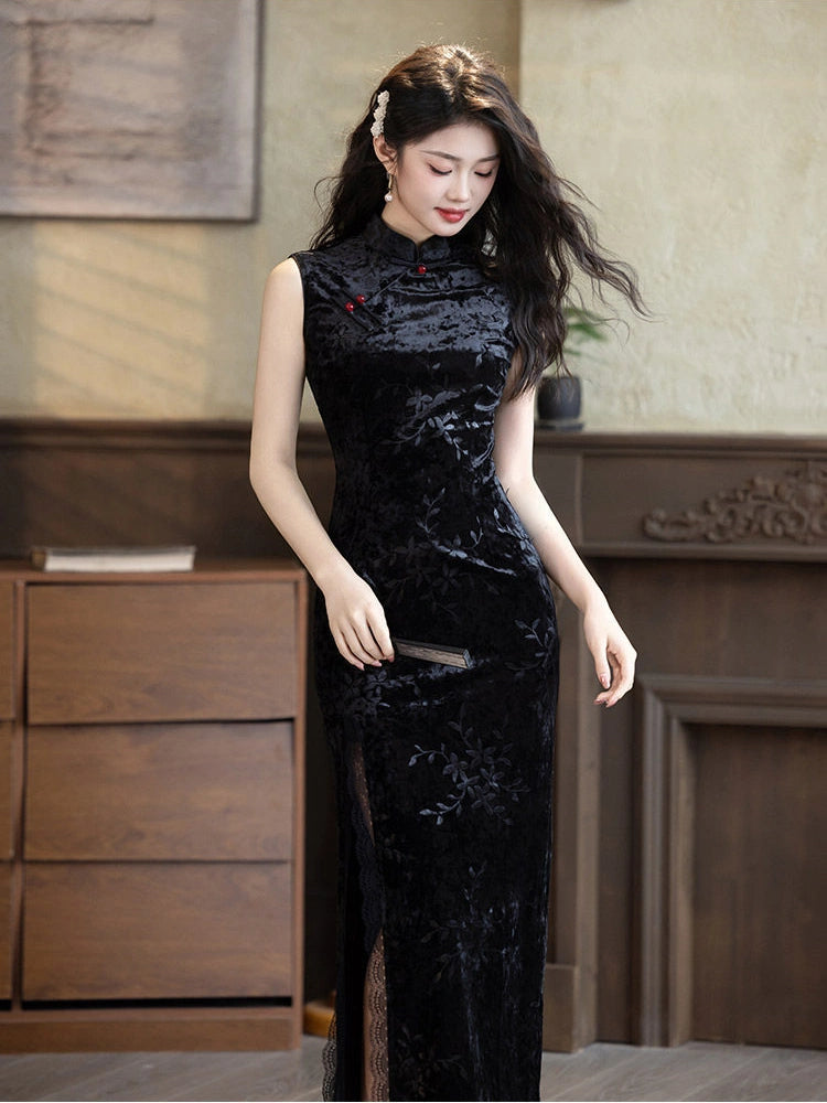 A woman wearing a sleeveless black velvet modern qipao cheongsam-style dress with floral embroidery and a high slit, standing in an indoor room with wooden furniture.