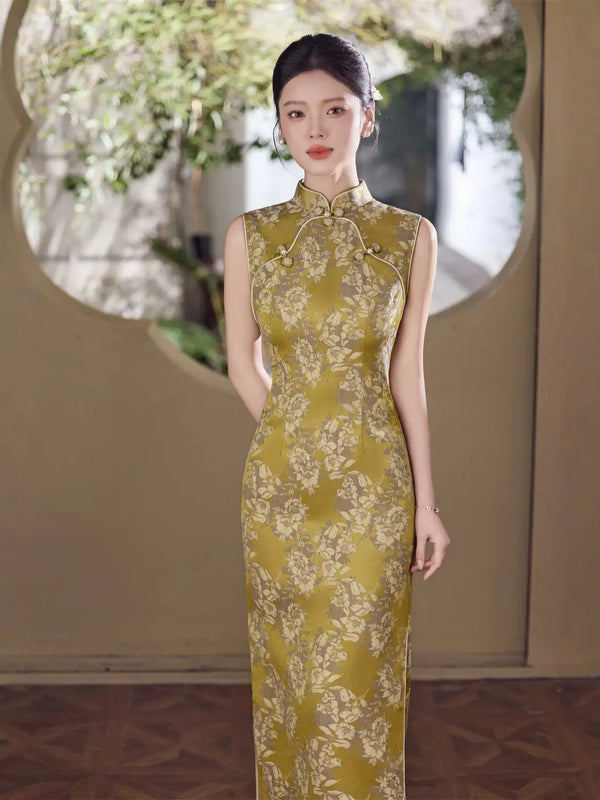 A woman stands indoors wearing a sleeveless midi cheongsam-style dress in olive yellow with a light floral pattern. The dress has a high collar and front button placket.