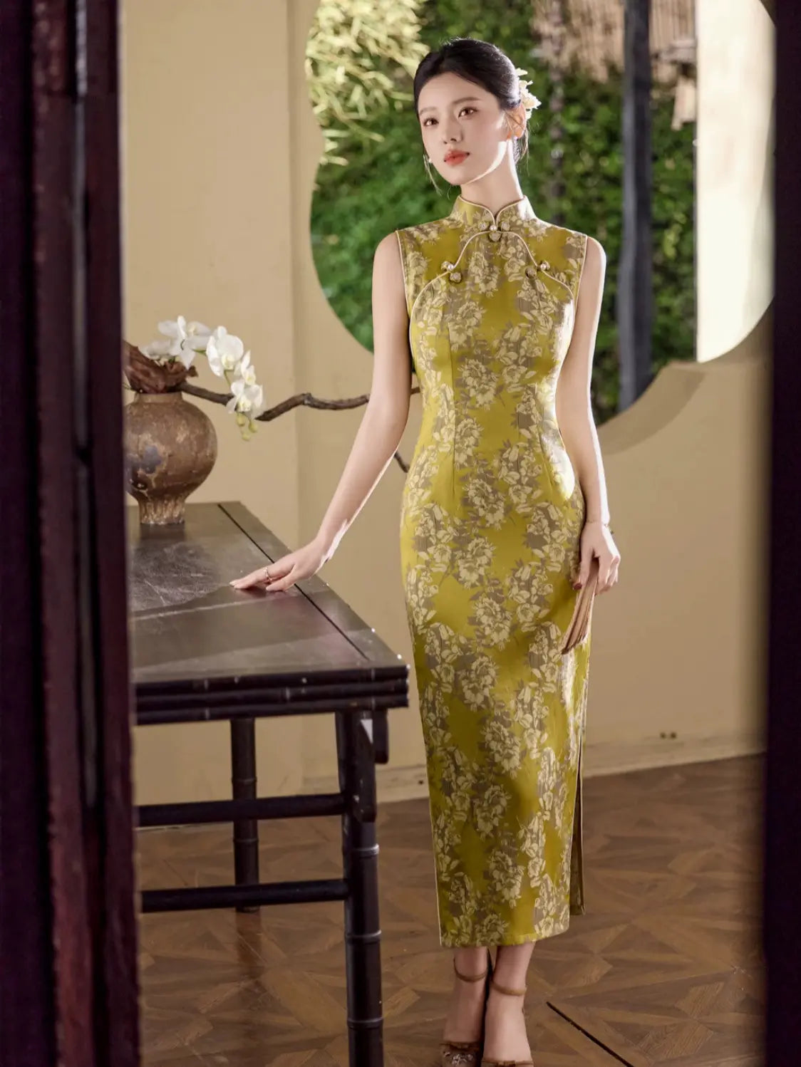 A woman wearing a sleeveless olive green and gold floral-patterned cheongsam (qipao) dress, standing in a traditional Chinese courtyard next to a wooden table.